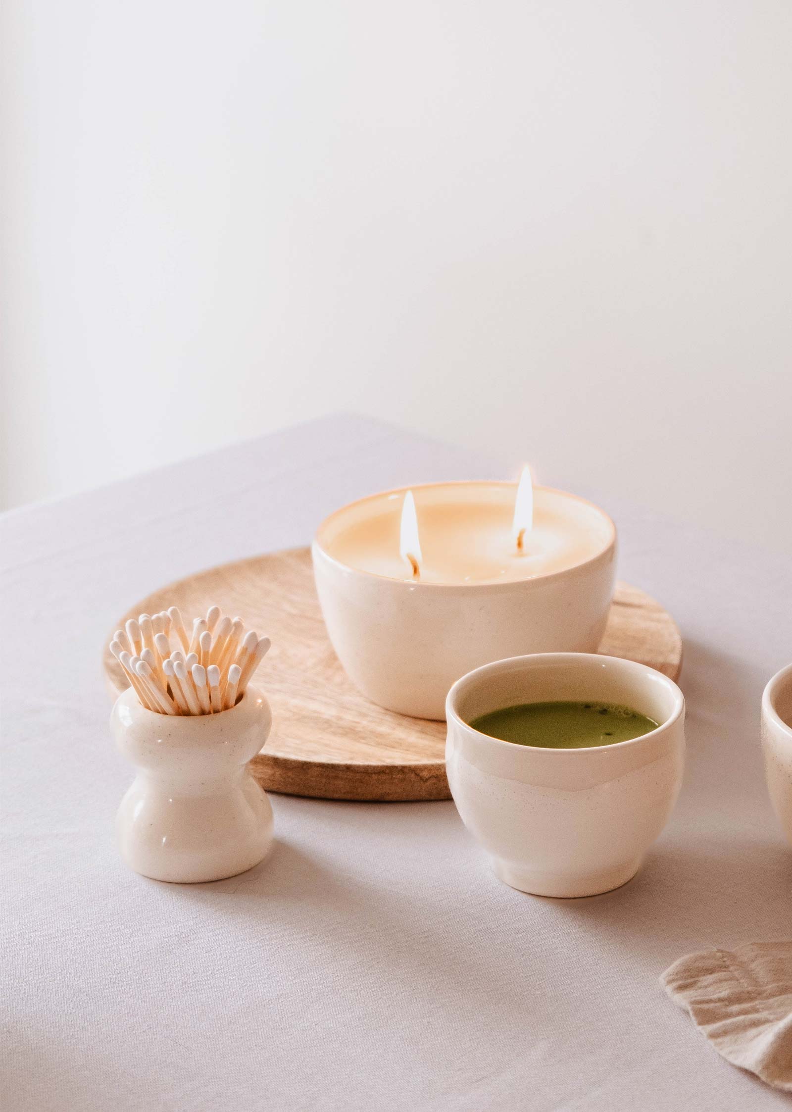 A lit candle in a ceramic holder adorns a wooden tray, accompanied by a small green beverage in a matching cup and an artisanal decor item—a white Mimi & August Petite Flamme Ceramic Match Pot, ideal for cotton swabs or matches.