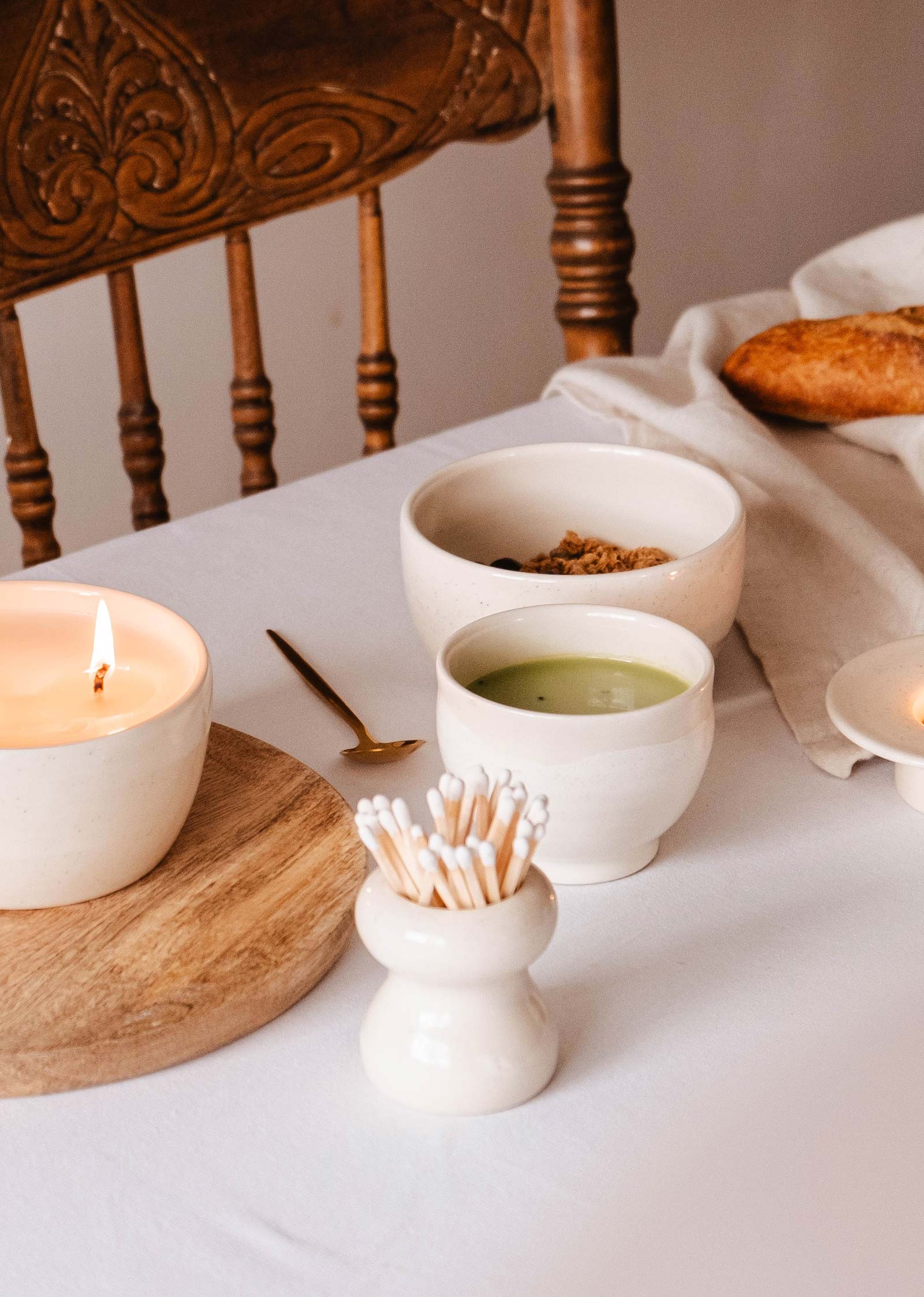 Une table avec une bougie allumée, le pot d'allumettes en céramique Mimi & August Petite Flamme, un bol de liquide vert, des céréales dans un autre bol, des cotons-tiges dans un support, une cuillère en laiton et du pain artisanal sur un tissu blanc, tout cela respire l'élégance artisanale et le charme multifonctionnel.