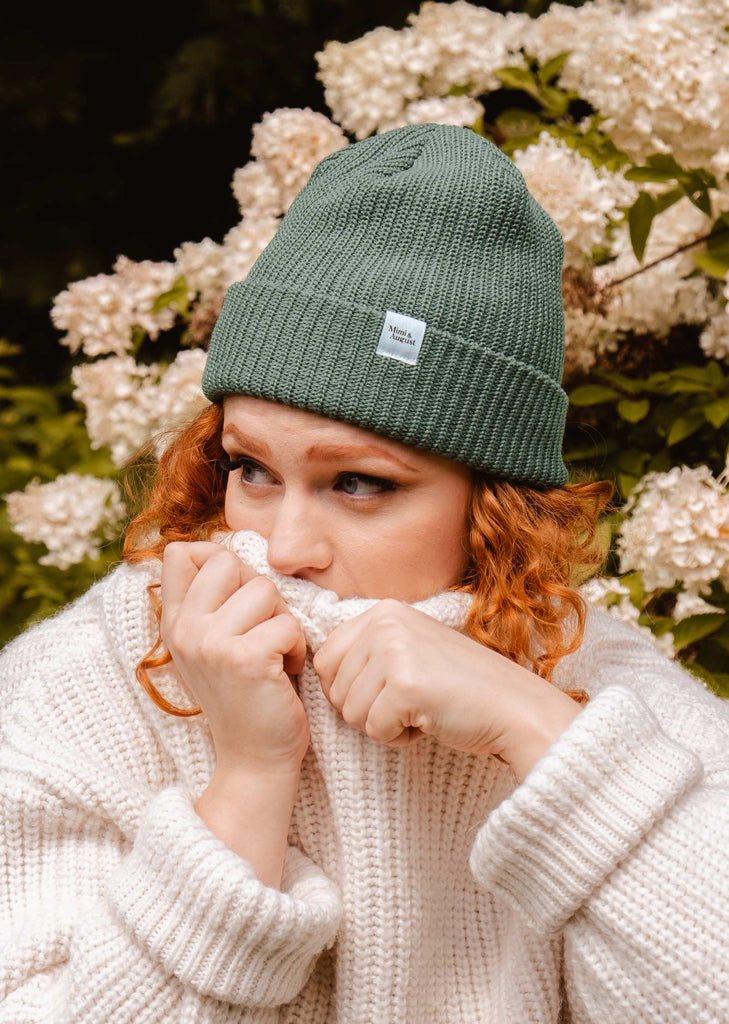 A woman wearing a mimi and august Pistachio Cuffed Beanie posing in front of flowers.