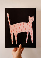 Hand holding a Playful Cat Art Print by Mimi & August featuring a stylized pink cat with light pink spots on a black background, all beautifully printed on recycled paper.