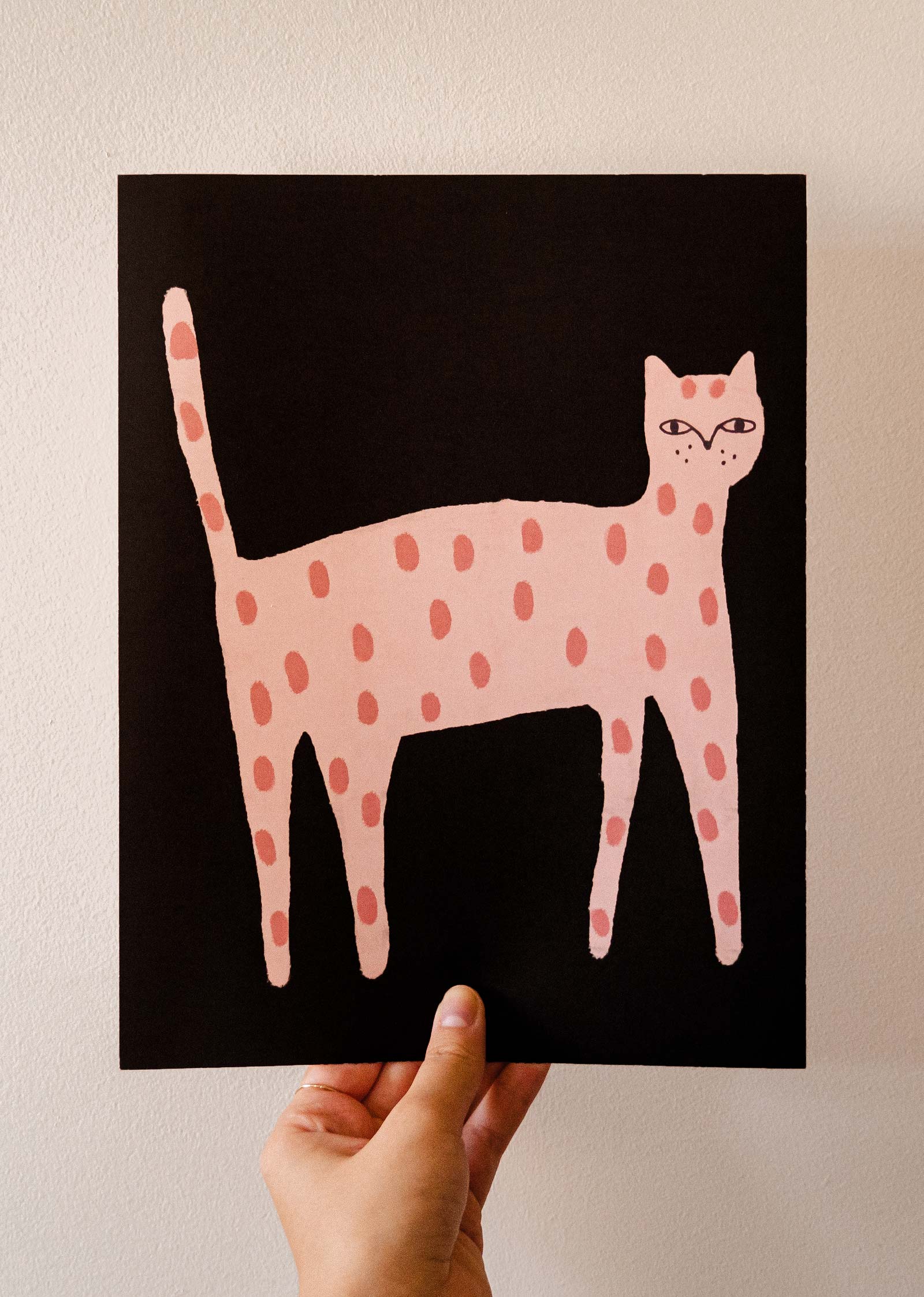 Hand holding a Playful Cat Art Print by Mimi & August featuring a stylized pink cat with light pink spots on a black background, all beautifully printed on recycled paper.