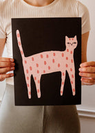 A person holds up a black poster featuring a pink, polka-dotted cat illustration. This Playful Cat Art Print by Mimi & August is crafted on recycled paper, adding an eco-friendly touch.