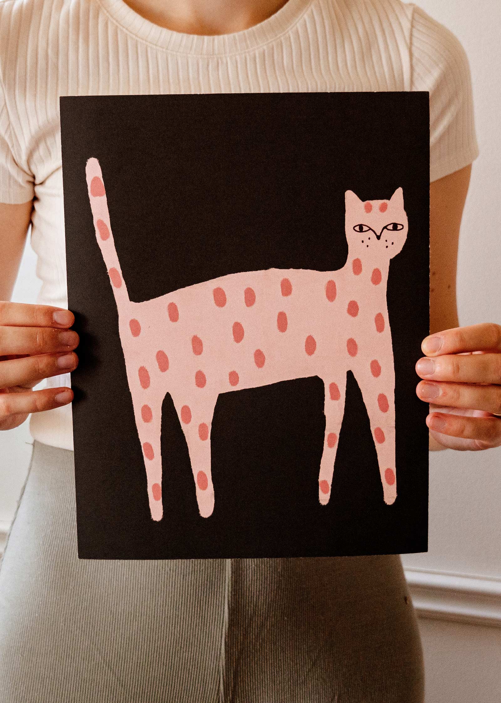 A person holds up a black poster featuring a pink, polka-dotted cat illustration. This Playful Cat Art Print by Mimi & August is crafted on recycled paper, adding an eco-friendly touch.