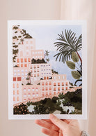 A hand holding up a Mimi & August Positano Art Print of Positano, an Italian coastal town with palm trees.