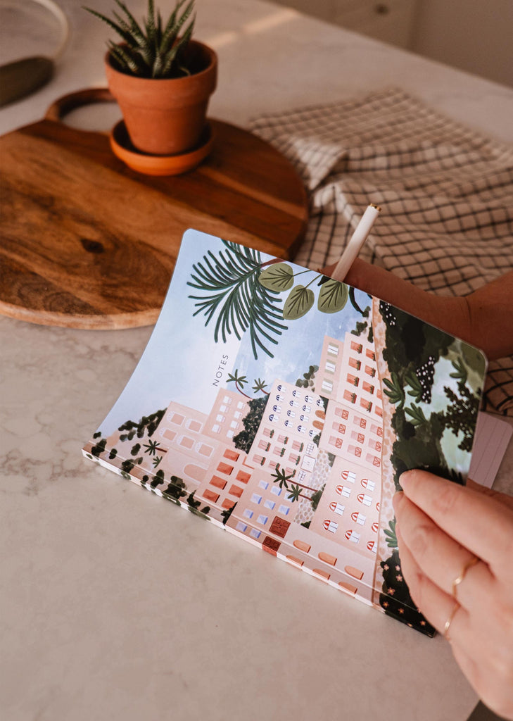 Une personne tenant un carnet de notes Mimi & August Positano représentant un paysage coloré avec un palmier, capturant l'essence d'une escapade relaxante.