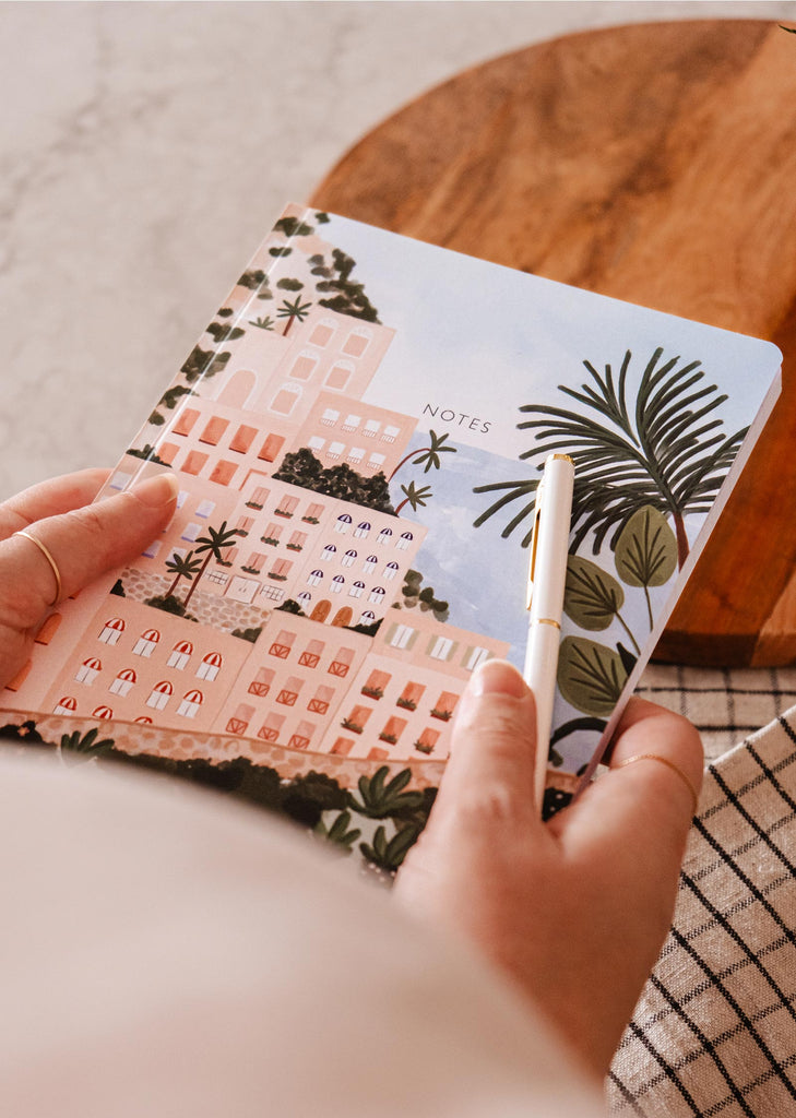 A person holding a Positano - Notebook from Mimi & August, with a palm tree on it, evoking a sense of getaway and blissful memories of Positano.