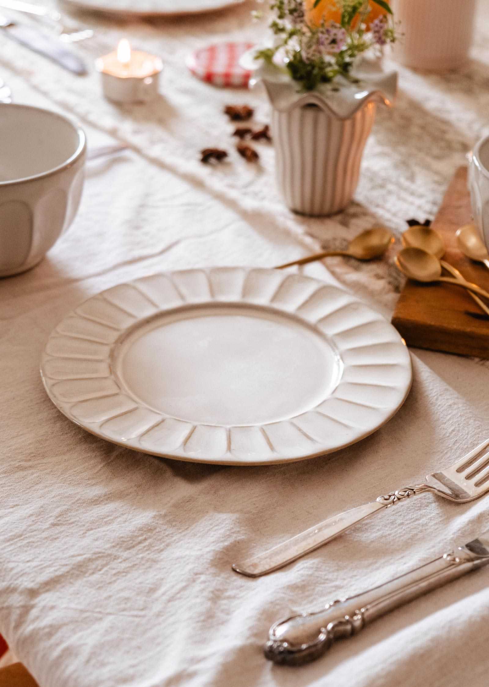 The handmade ceramic plate by Mimi & August from our ceramic breakfast collection sits on a linen-covered table, accompanied by a fork, knife, gold spoons, flowers in a vase, and a lit tea candle nearby.