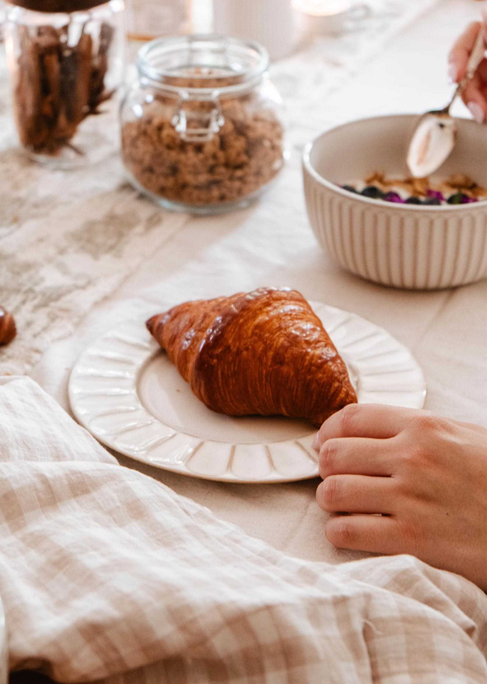 A hand reaches for a croissant on The handmade ceramic plate by Mimi & August, with a bowl of granola and a jar of cereal from the ceramic breakfast collection resting on a light-colored tablecloth in the background.