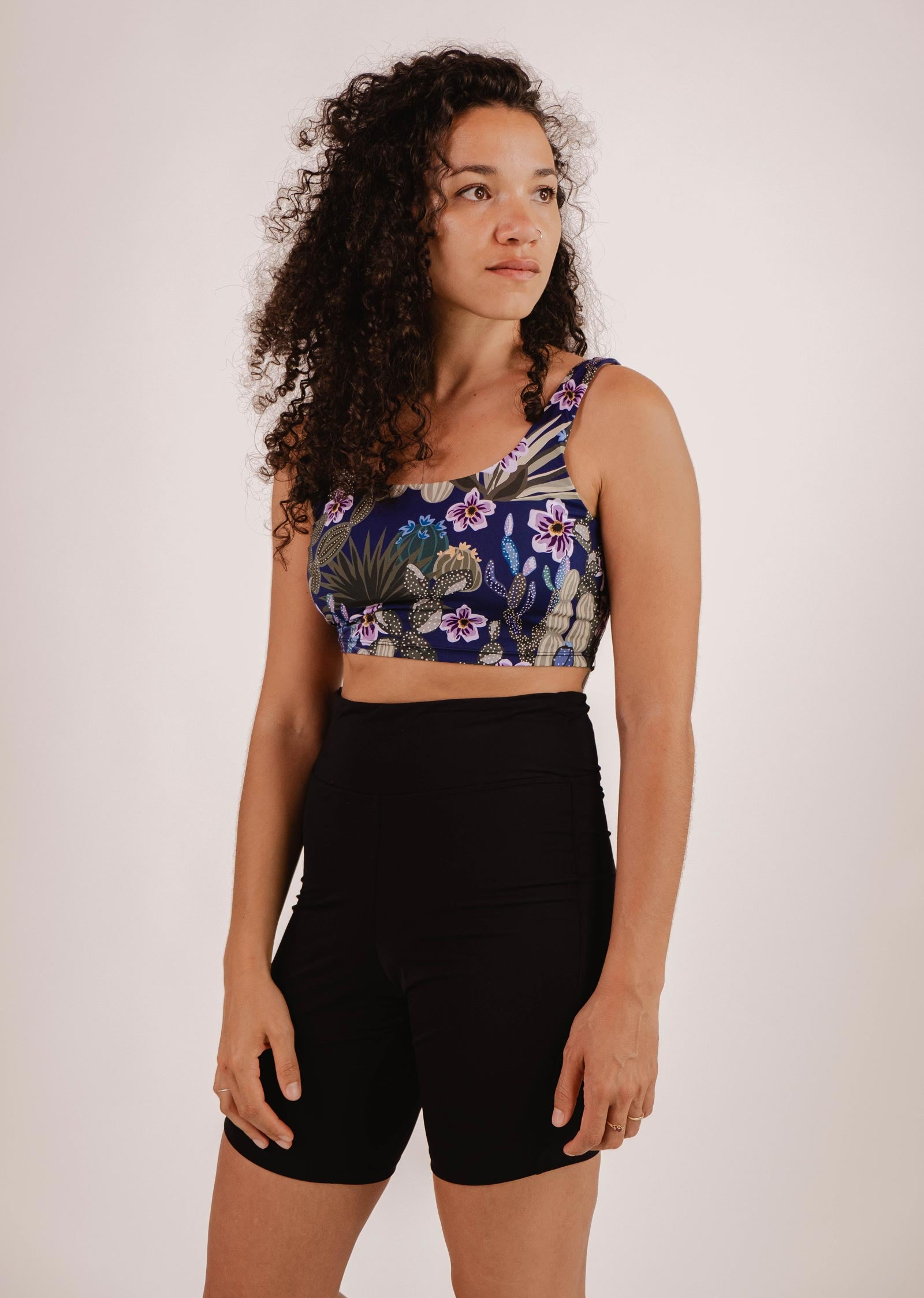 Une personne aux cheveux bouclés regarde hors champ, vêtue d'un crop top à fleurs et d'un short de vélo de bain taille haute Riviera Noir de Mimi & August  sur un fond uni.