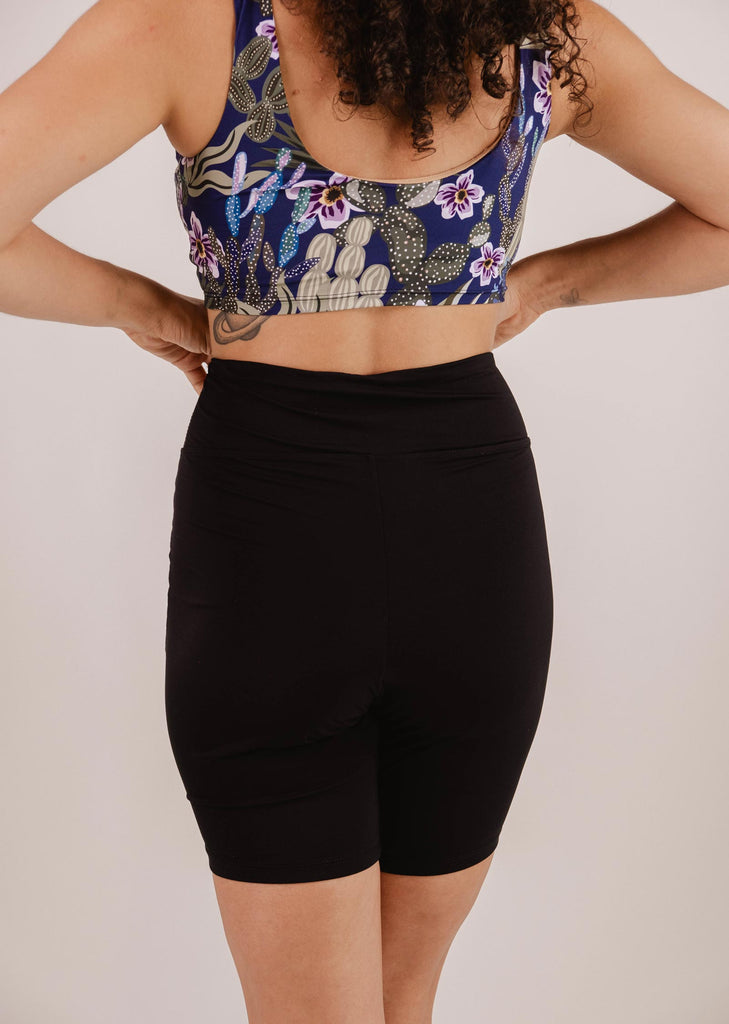 A person in a floral sports bra and black Mimi & August Riviera Black High Waist Swim Bike Short made from Econyl swimsuit fabric is standing with their hands on their hips, facing away.