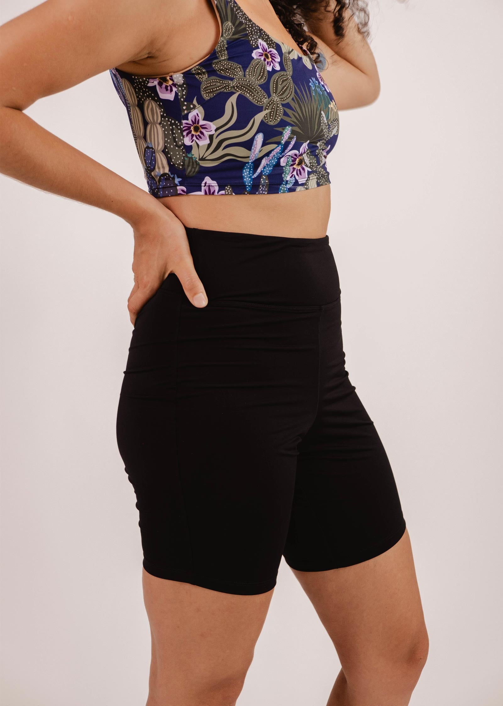 Une personne portant un soutien-gorge de sport à motifs floraux et le short de vélo de bain Riviera Black High Waist Swim Bike de Mimi & August en tissu de maillot de bain Econyl se tient debout, les mains sur les hanches.