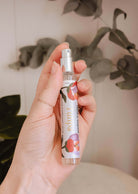 Une main tient un flacon de spray d'ambiance Mimi & August - Tulipa avec un motif floral, évoquant de délicates notes florales, sur fond de feuilles vertes et d'un mur de couleur claire.