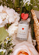 Un flacon de spray d'ambiance Mimi & August - Amore avec un bouchon en forme de cœur rouge est posé parmi des fleurs blanches et roses, créant une atmosphère romantique dans un panier tressé.