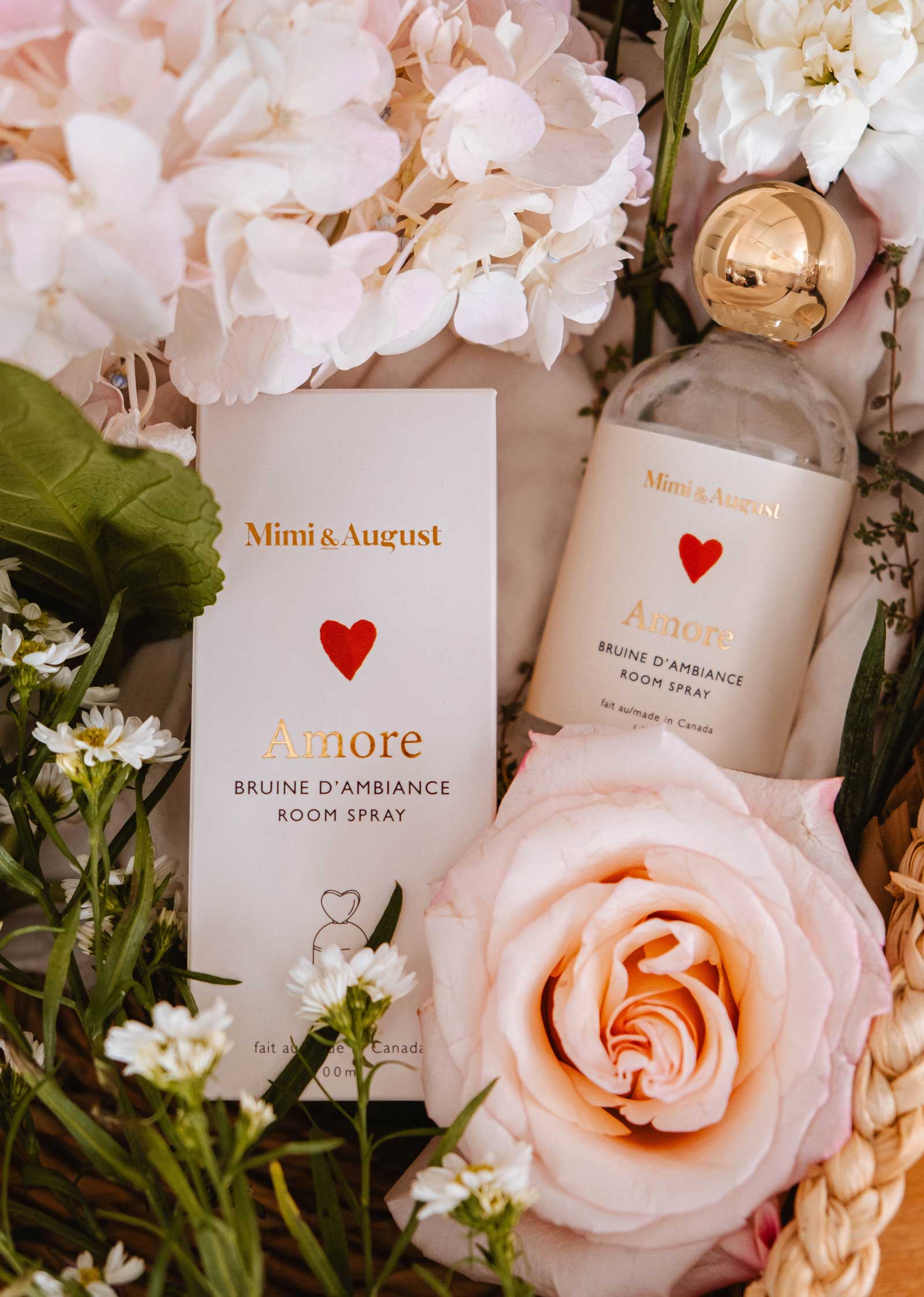 The Room spray - Amore by Mimi & August, shown with pink and white flowers and lush greenery, creates a romantic vibe with its delicate floral scent.