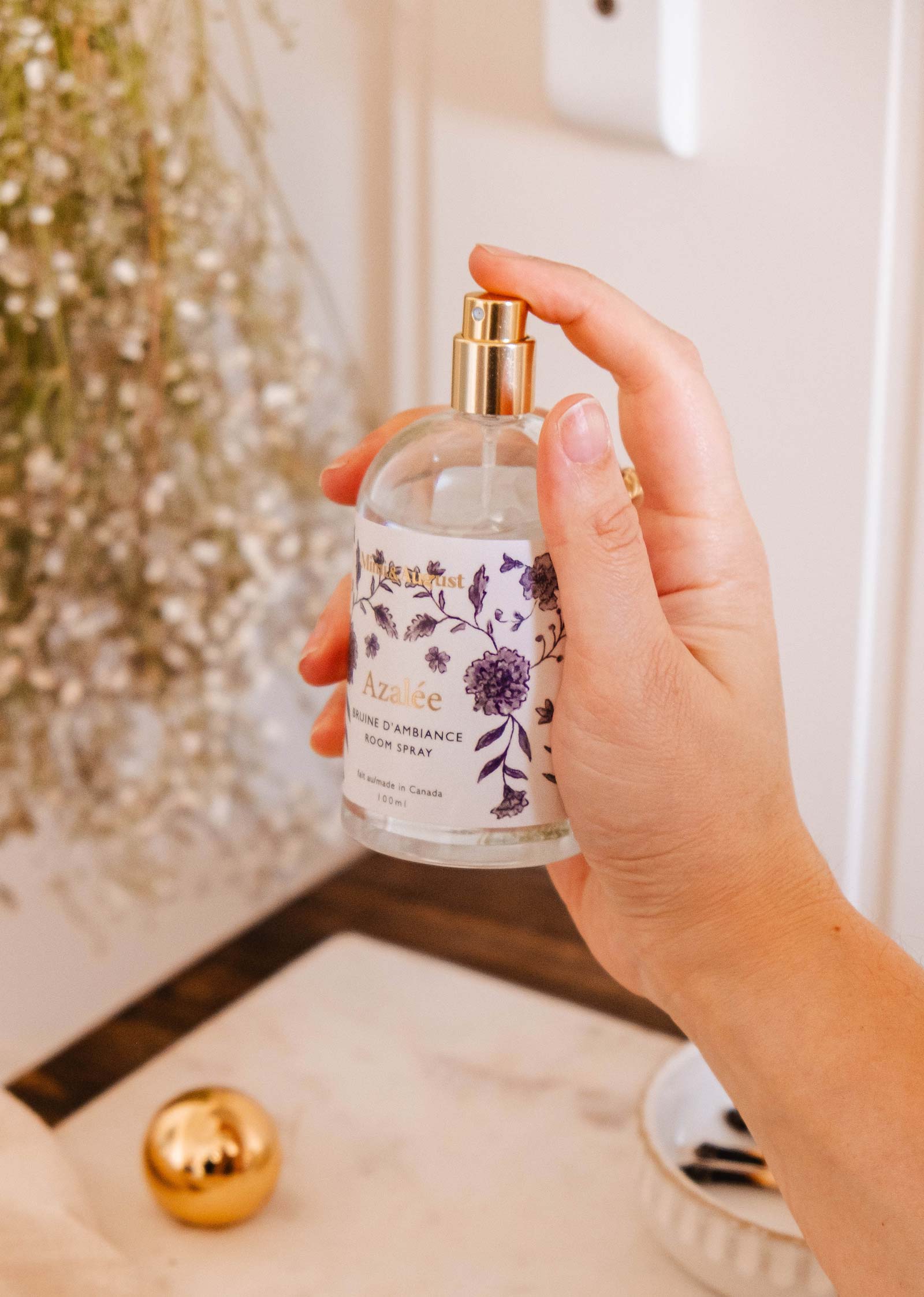 A hand holds a Mimi & August Room spray - Azalée bottle, with a floral label, near a bowl containing black stones and a golden sphere. In the background, white flowers that resemble jasmine blooms hang gracefully.