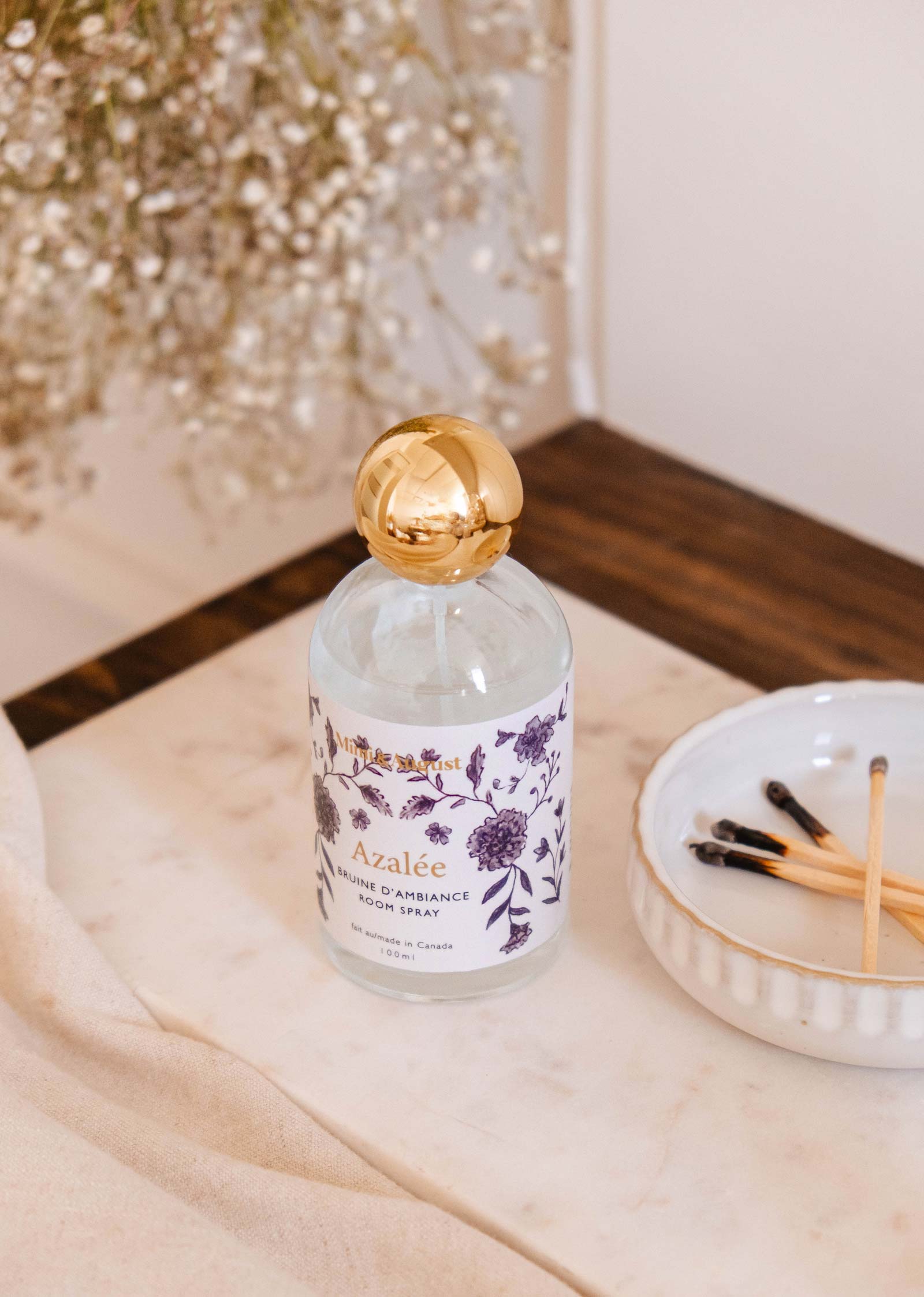 A Mimi & August Azalée Room Spray bottle with a floral label and gold cap sits on marble beside matchsticks, releasing jasmine hints. Hanging flowers enhance the serene ambiance.