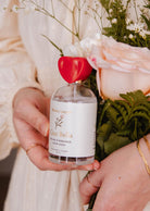 Une personne tient la bruine d'ambiance- Ciao Bella de Mimi & August, avec son bouchon en forme de cœur rouge, niché à côté d'un bouquet de roses roses et de feuillage vert. Le parfum floral et boisé se mêle subtilement à des arômes d'agrumes frais, évoquant une senteur invitante qui rappelle les jardins en fleurs.