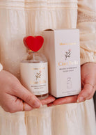 Une personne tient un spray d'ambiance Ciao Bella de Mimi & August, doté d'un bouchon en forme de cœur rouge et d'une boîte assortie, qui émet un parfum floral et boisé.