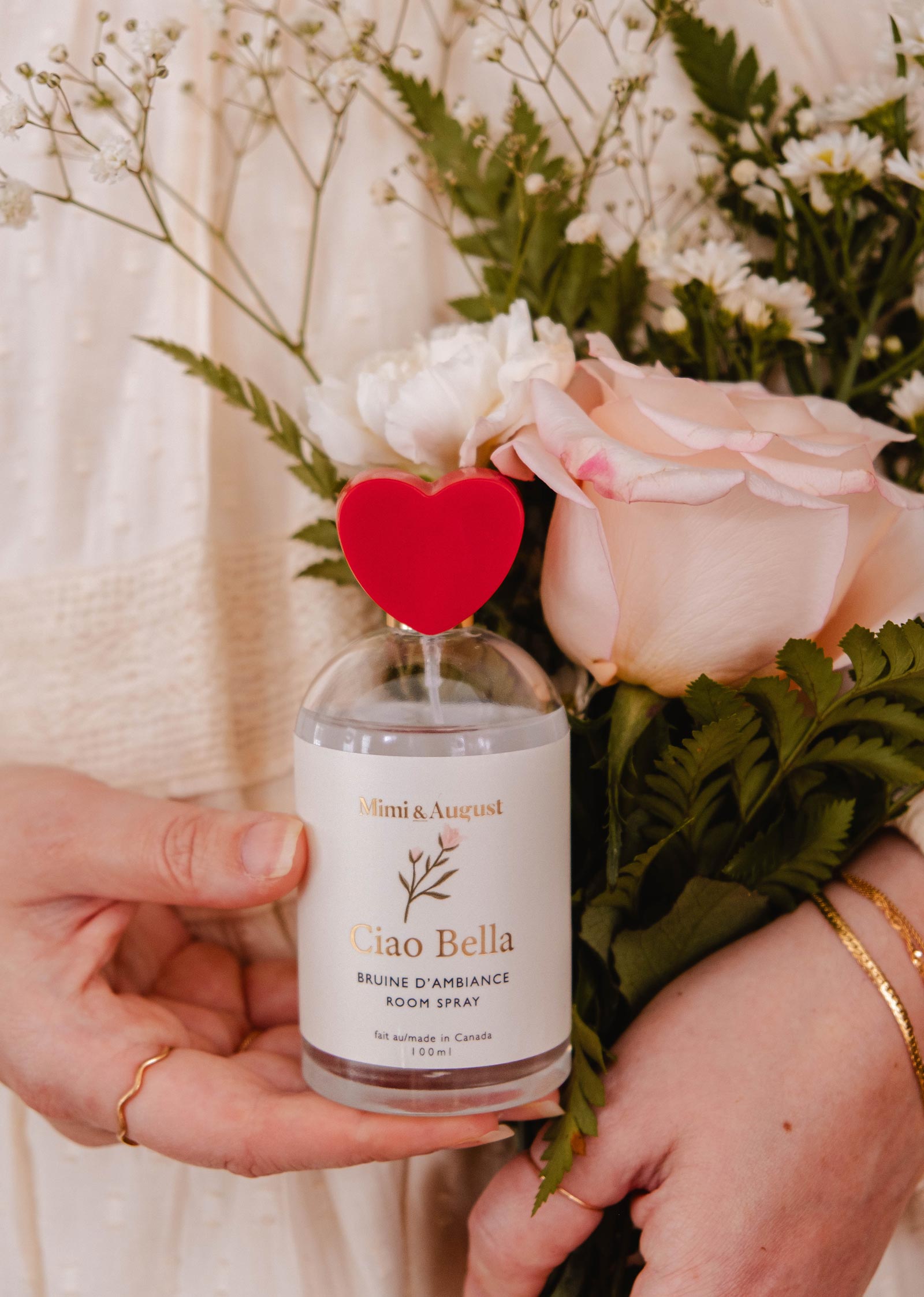 Une main tient le vaporisateur d'ambiance Mimi & August "Ciao Bella", doté d'un bouchon en forme de cœur rouge. Entourée de roses roses et d'un feuillage vert luxuriant, la scène reflète son arôme floral rafraîchissant.