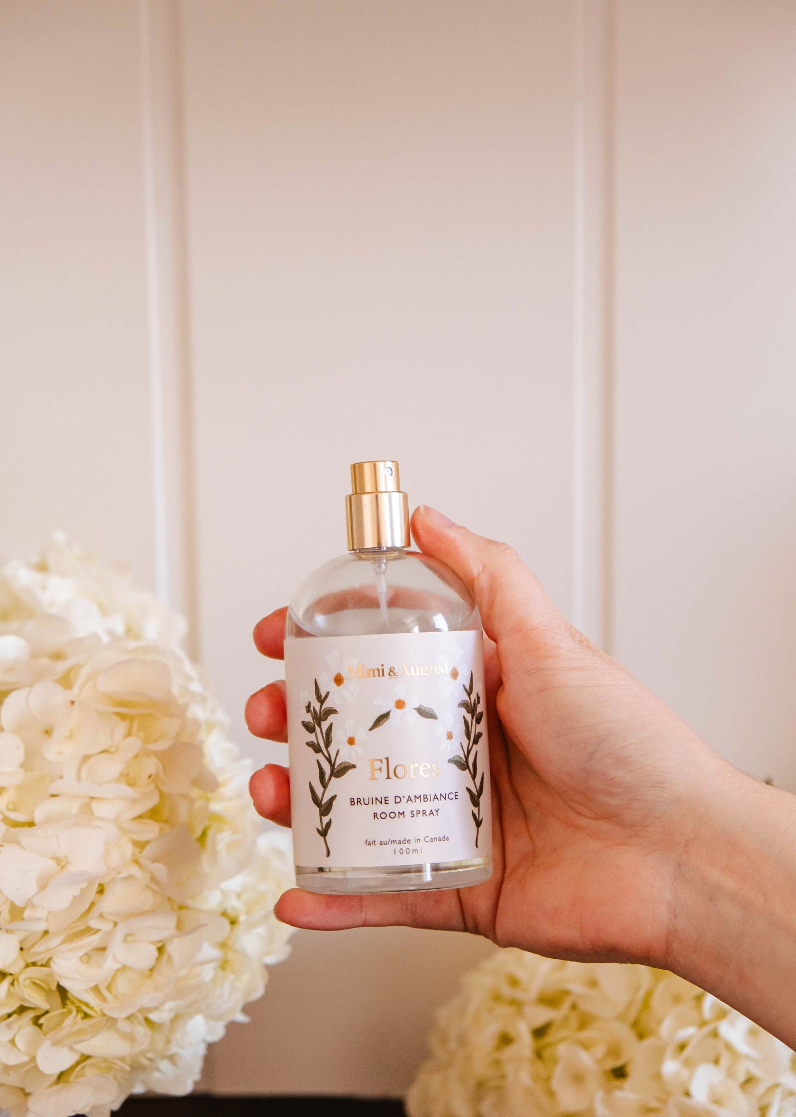 A hand holds Mimi & August's "Room Spray - Flores" surrounded by white hydrangeas, promising a floral fragrance that refreshes the atmosphere against a serene, light backdrop.
