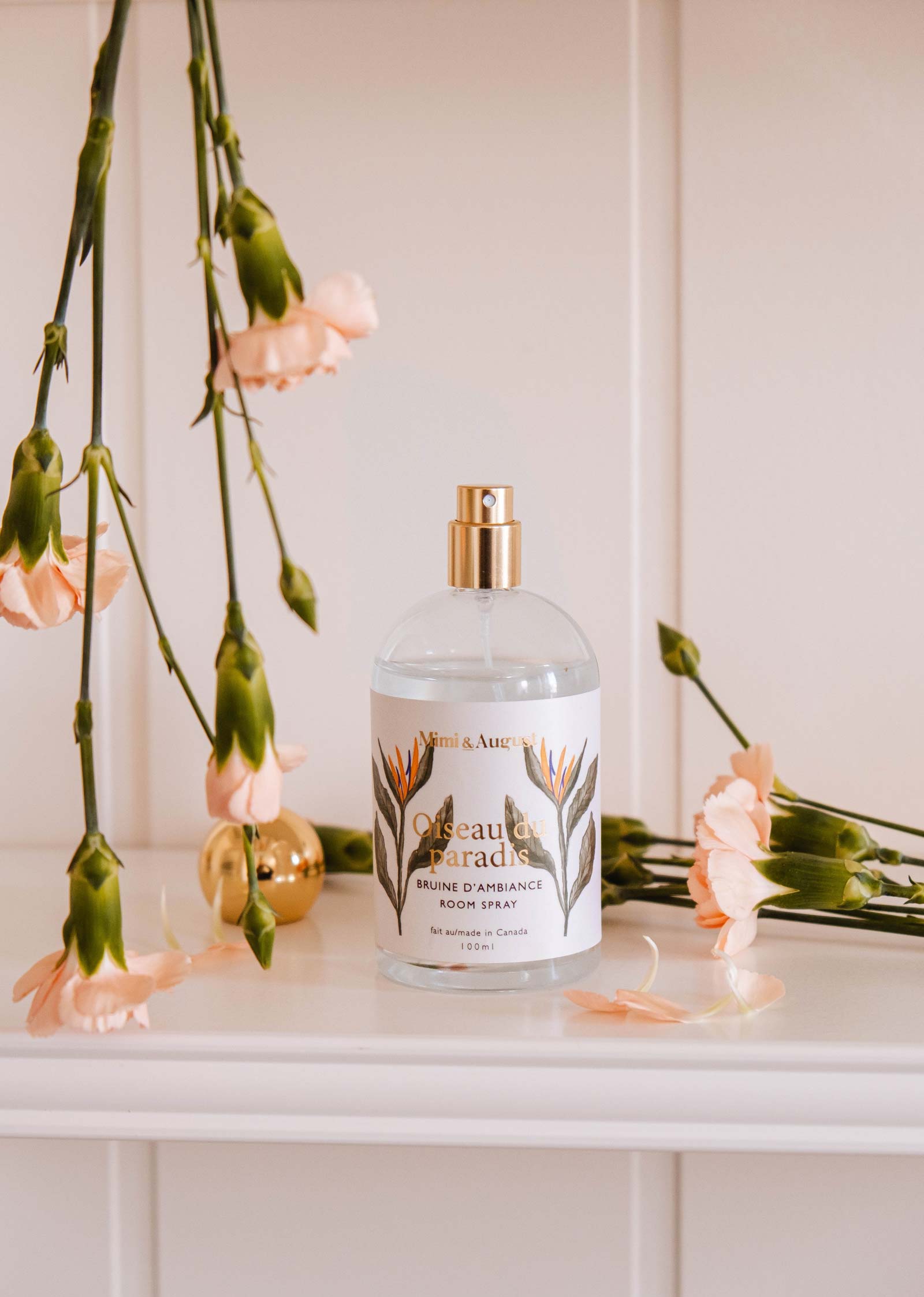 A Mimi & August Room spray - Oiseau du paradis, in a clear glass bottle with a floral label, sits on a shelf amidst light pink carnations, releasing delicate citrus and tropical fruit aromas.
