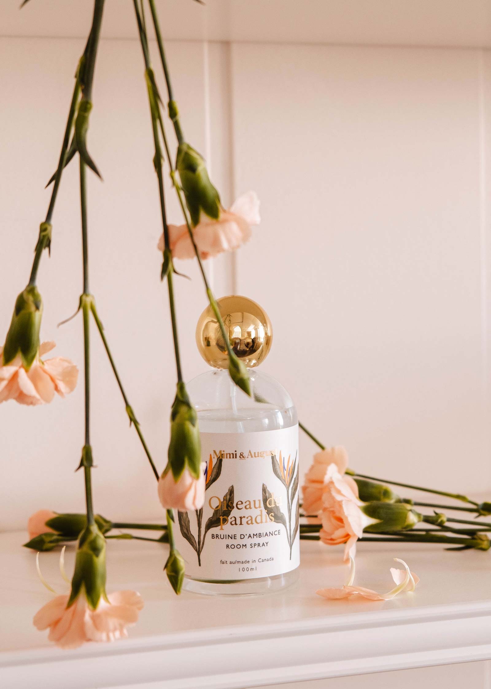 On a white surface, amid peach carnations, rests a Mimi & August Room Spray - Oiseau du paradis with a gold cap, emitting a citrusy and fruity tropical mist.