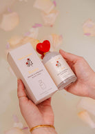 Hands holding the Mimi & August Room Spray - Rosea, exuding floral elegance with a hint of citrus zest. The chic packaging, complemented by a red heart cap and floating petals, adds a delicate charm to the background.