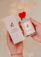 Hands holding a Mimi & August Room Spray - Rosea bottle, featuring a heart-shaped cap, exuding floral elegance, beside packaging adorned with roses and citrus zest hints.