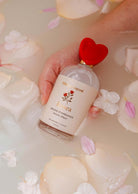 A hand holding a Mimi & August Room Spray - Rosea with a red heart-shaped cap, surrounded by rose petals in water, exudes floral elegance with a hint of citrus zest.