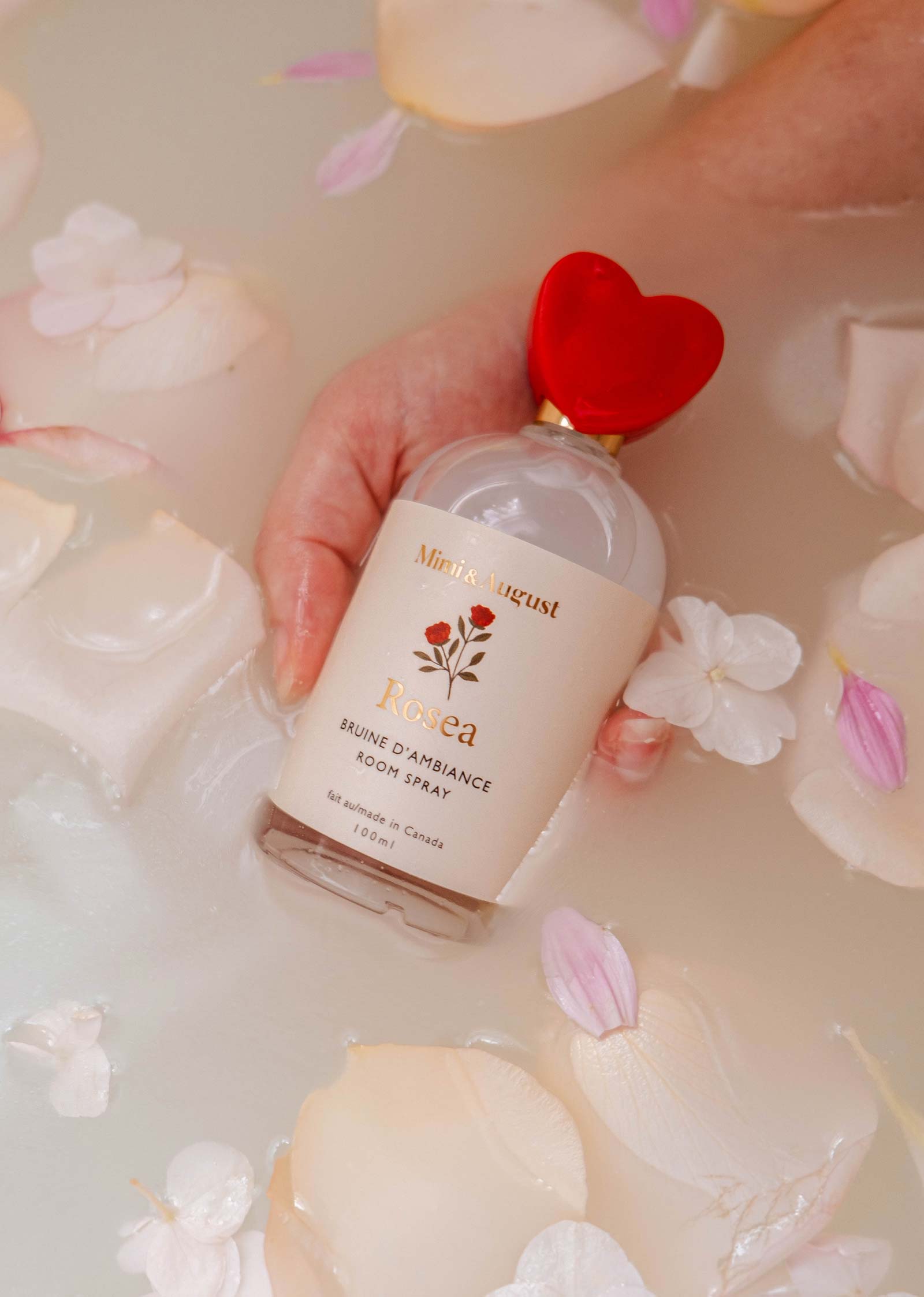 A hand holding a Mimi & August Room Spray - Rosea with a red heart-shaped cap, surrounded by rose petals in water, exudes floral elegance with a hint of citrus zest.