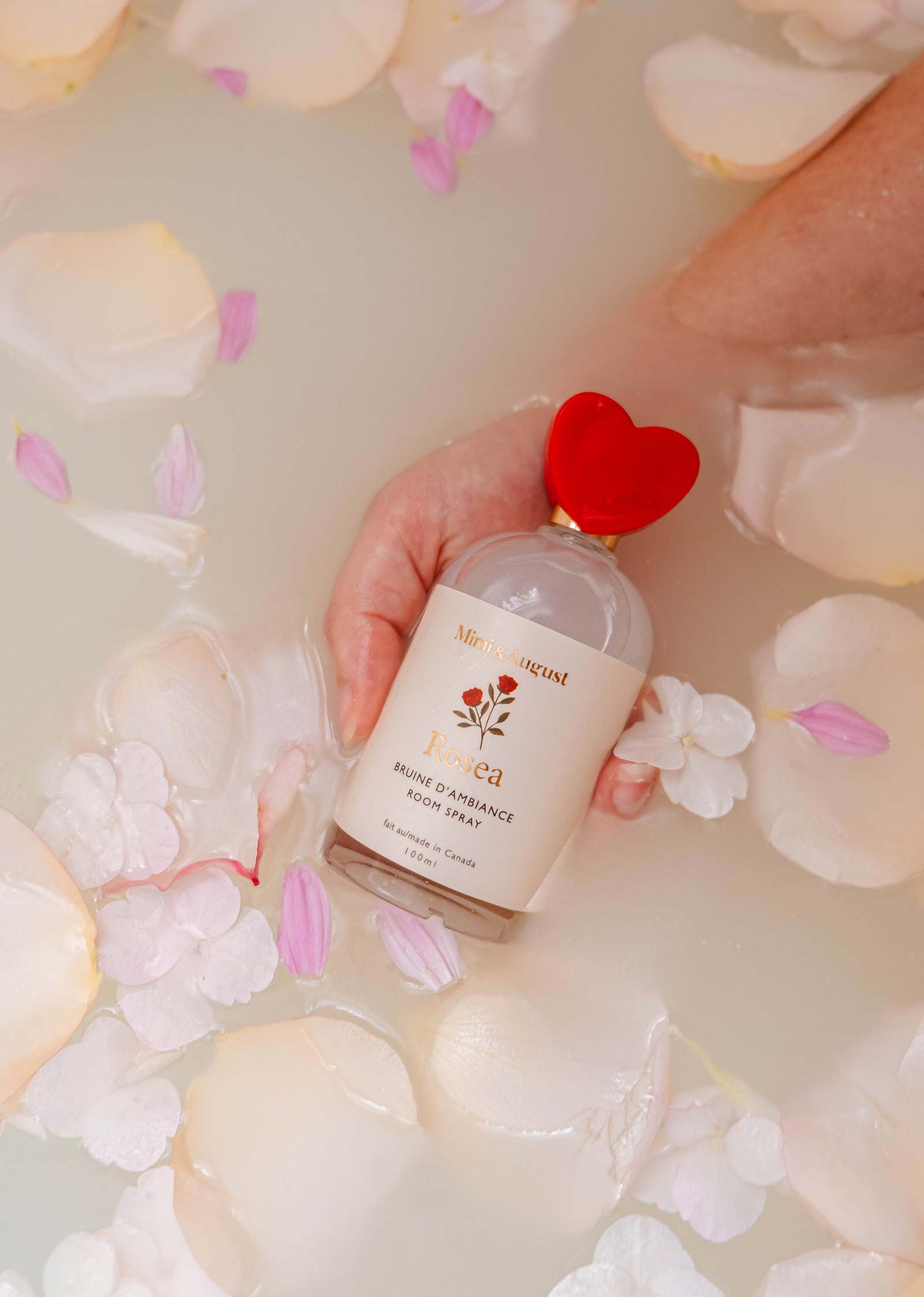 A hand holds a bottle of Mimi & August's Room Spray - Rosea with a red heart cap in a milky bath surrounded by pink and white flower petals, exuding floral elegance.