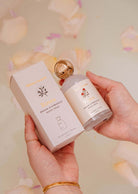 A person holds a box and bottle of "Room spray - Rosea" by Mimi & August, a citrus floral room spray, surrounded by flower petals in soft, warm lighting.