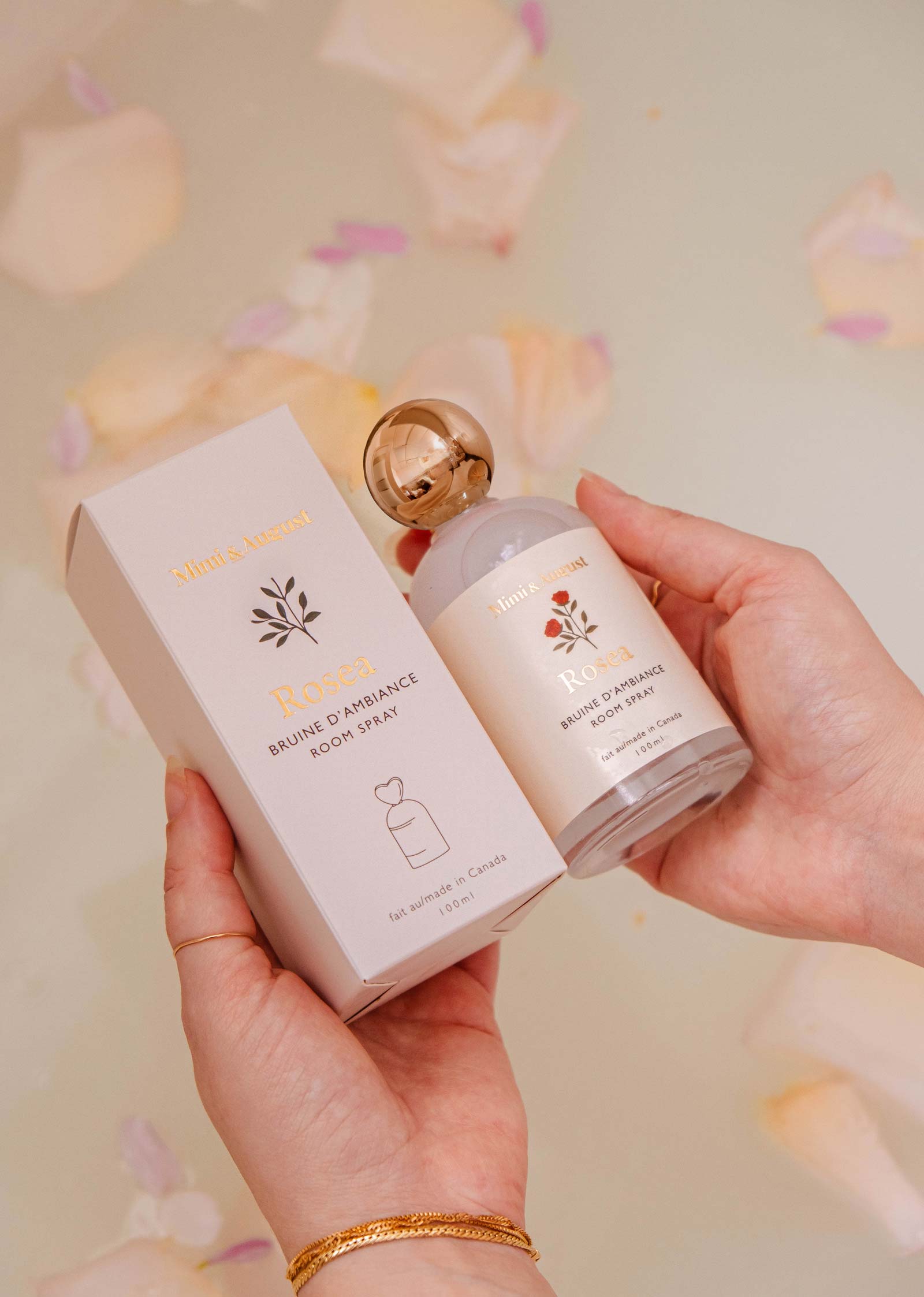 A person holds a box and bottle of "Room spray - Rosea" by Mimi & August, a citrus floral room spray, surrounded by flower petals in soft, warm lighting.