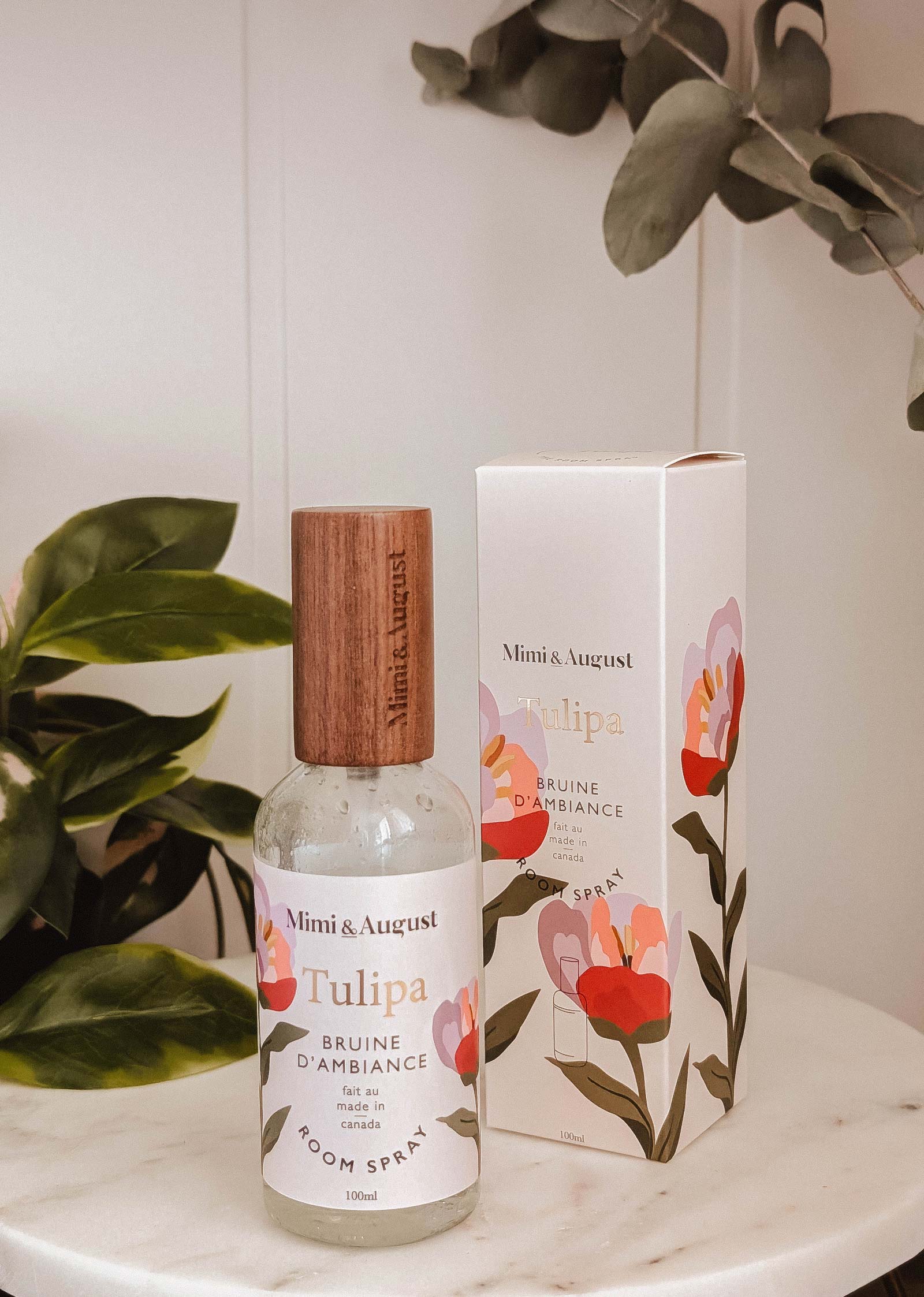 A bottle of Mimi & August Room Spray - Tulipa with a wooden cap sits by its floral-patterned box on a white surface, surrounded by green leaves. Notes of wildberries and fresh florals linger in the air.