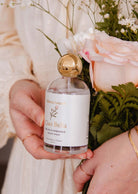 Someone holds a bottle of Mimi & August's Room Spray - Ciao Bella next to soft pink roses and greenery, showcasing this elegant home fragrance with its delicate floral aroma.