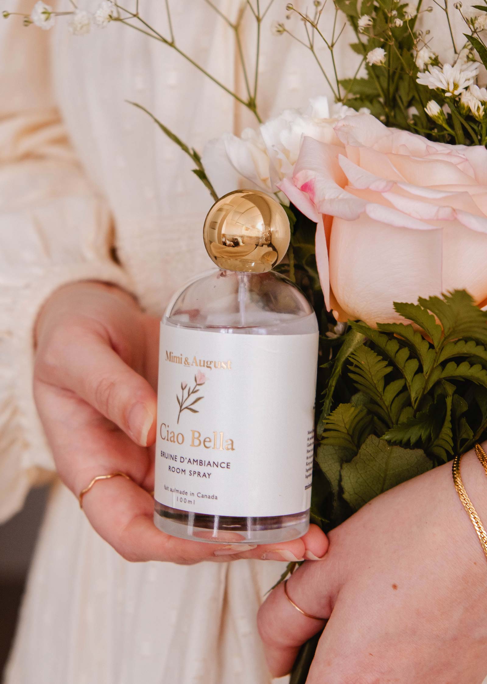 Someone holds a bottle of Mimi & August's Room Spray - Ciao Bella next to soft pink roses and greenery, showcasing this elegant home fragrance with its delicate floral aroma.
