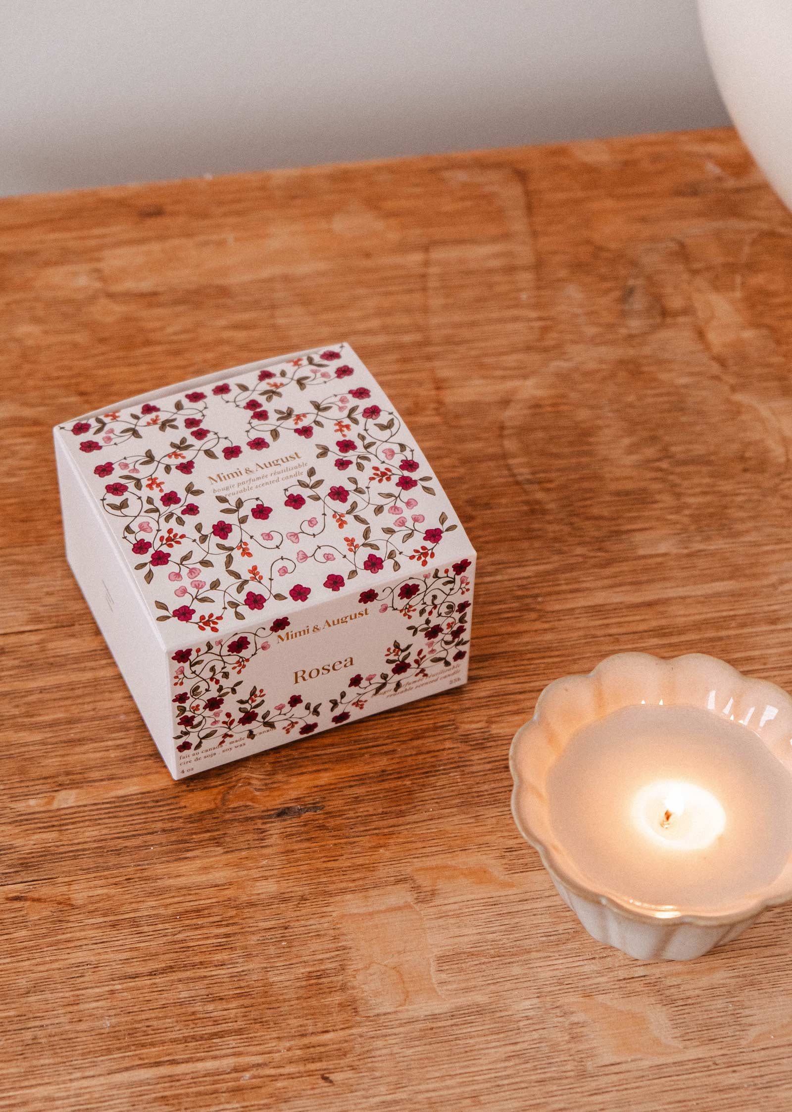 A floral-patterned Rosea - Reusable Candle by Mimi & August sits on a wooden surface in a white, scalloped-edge holder, radiating an uplifting scent.