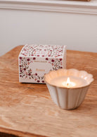 The hand-poured Rosea - Reusable Candle by Mimi & August glows in a scalloped holder on wood, topped with uplifting scent beside its floral-patterned box.