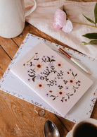 The Rosie notebook by Mimi & August sits on a planner with a pen and pink flower, all made from recycled paper, atop a wooden table alongside a coffee cup and pitcher.