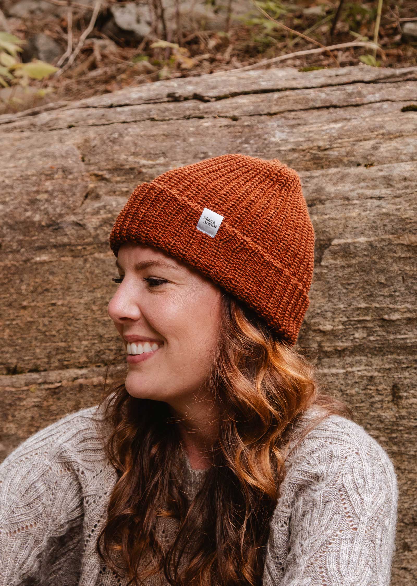 Une femme aux longs cheveux bouclés, portant un bonnet Rust Chunky Beanie de Mimi & August et un pull gris, sourit alors qu'elle est assise en plein air sur un fond rocheux. Le bonnet, son accessoire d'hiver préféré, ajoute une touche de chaleur à la scène.