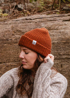 A person wearing their cozy favorite, the Rust Chunky Beanie by Mimi & August, and a beige sweater is standing outdoors against a rocky background.