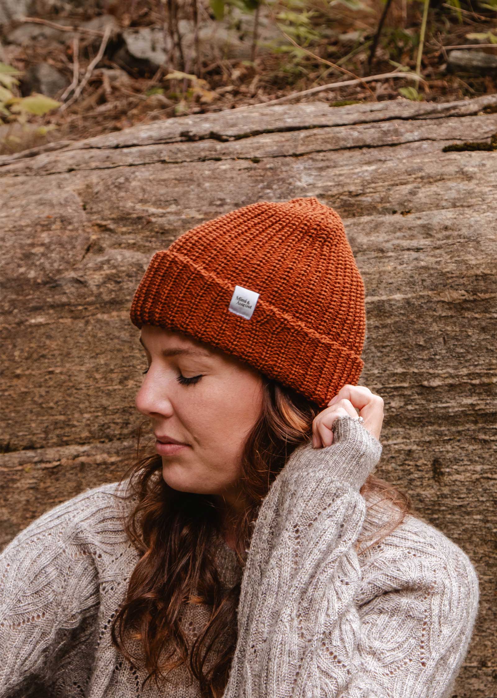 Une personne portant son bonnet préféré, le Rust Chunky Beanie de Mimi & August, et un pull beige se tient à l'extérieur sur un fond rocheux.