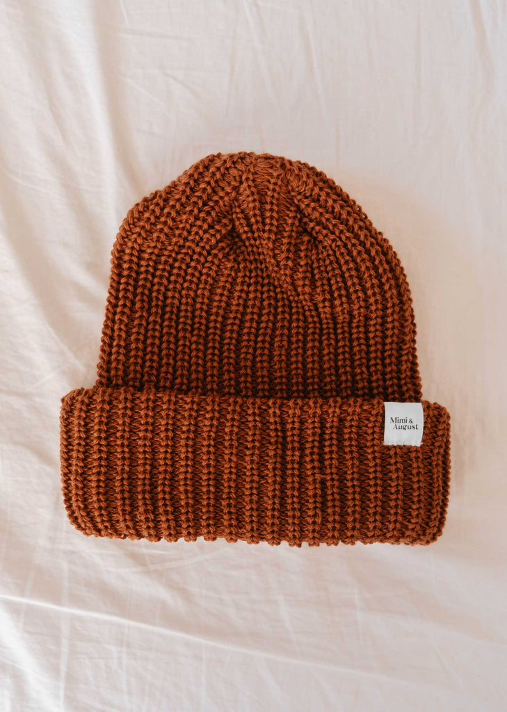 A folded Rust Chunky Beanie by Mimi & August, in a warm rust color with a cozy texture and adorned with a small white label on the cuff, resting on a light-colored fabric background.