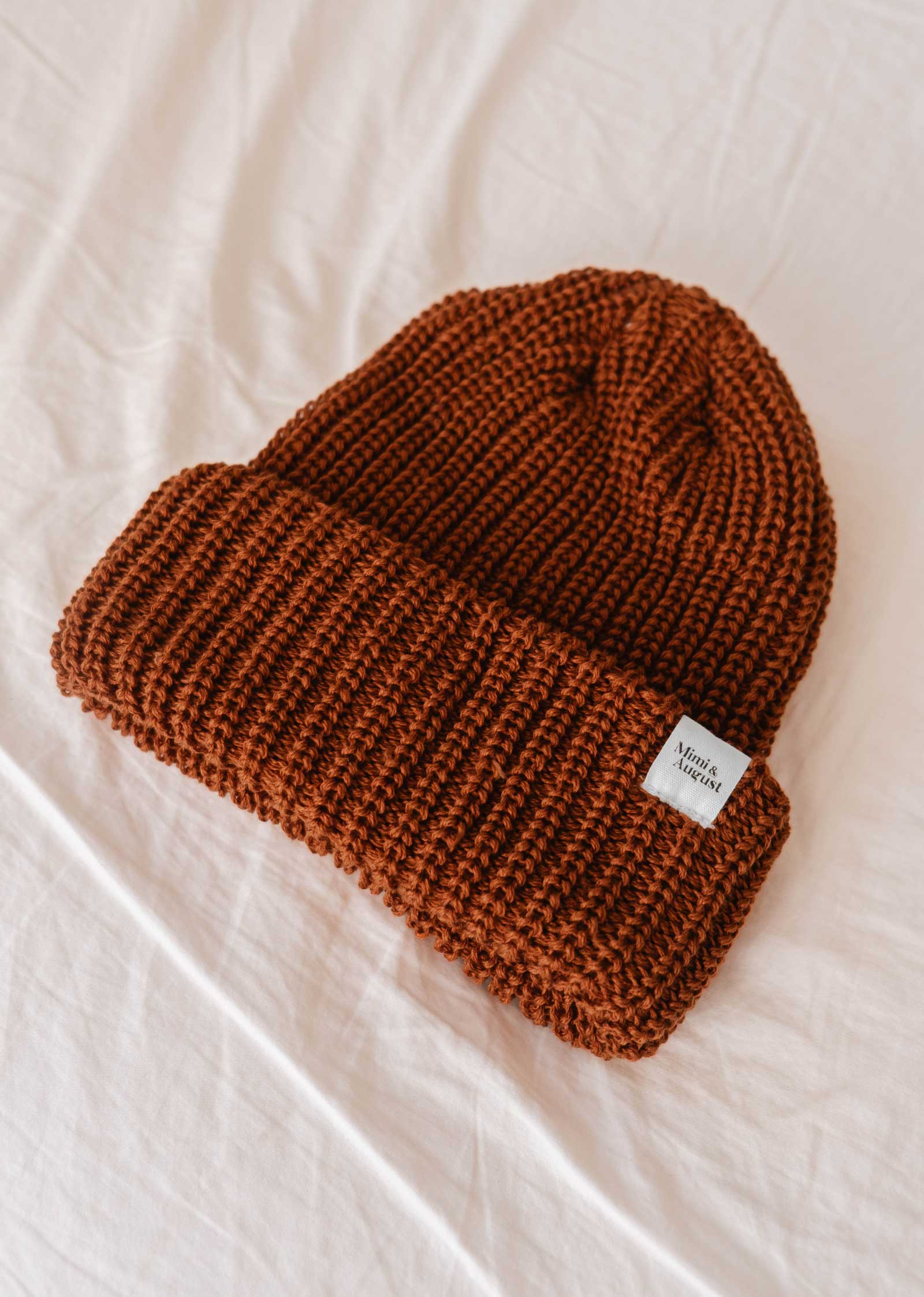 Le bonnet Rust Chunky Beanie de Mimi & August, doté d'un bord plié et d'une petite étiquette sur un côté, repose sur une surface en tissu léger ; ce favori douillet deviendra à coup sûr votre basique de l'hiver.