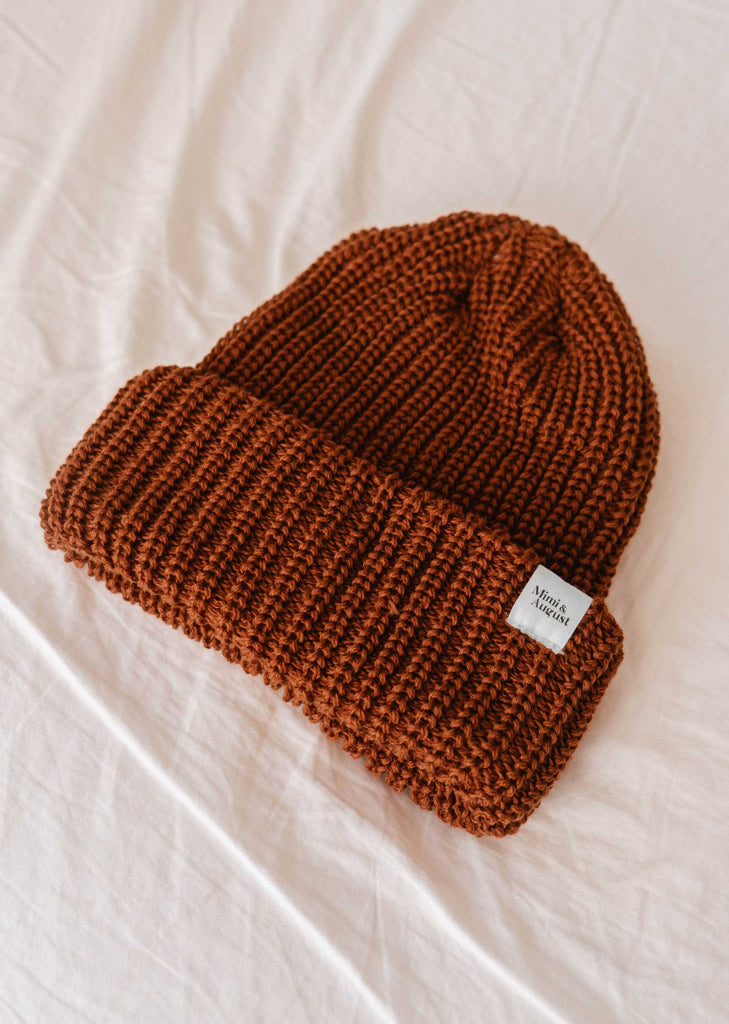 Le bonnet Rust Chunky Beanie de Mimi & August, doté d'un bord plié et d'une petite étiquette sur un côté, repose sur une surface en tissu léger ; ce favori douillet deviendra à coup sûr votre basique de l'hiver.