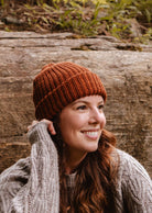 A woman wearing a cozy favorite, the Mimi & August Rust Chunky Beanie and a gray sweater, smiles while sitting outdoors near a large rock.