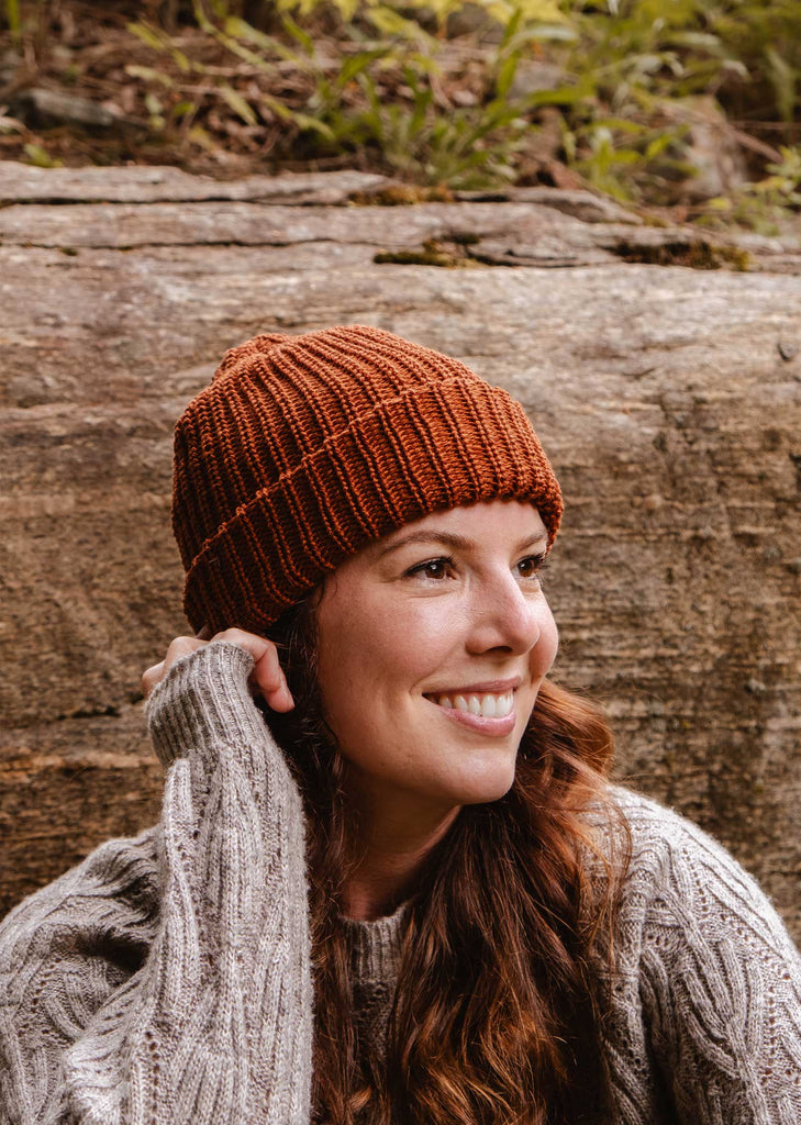 Une femme portant un de ses bonnets préférés, le Mimi & August Rust Chunky Beanie, et un pull gris, sourit alors qu'elle est assise à l'extérieur, près d'un gros rocher.