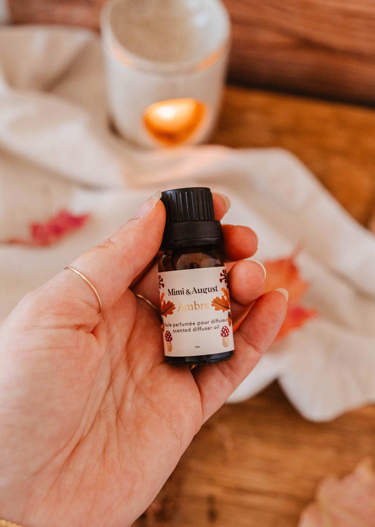 A hand holds a small bottle of "Scented Oil - Ambre" by Mimi & August, with a blurred background showcasing wooden textures, tropical foliage, and an illuminated candle holder.