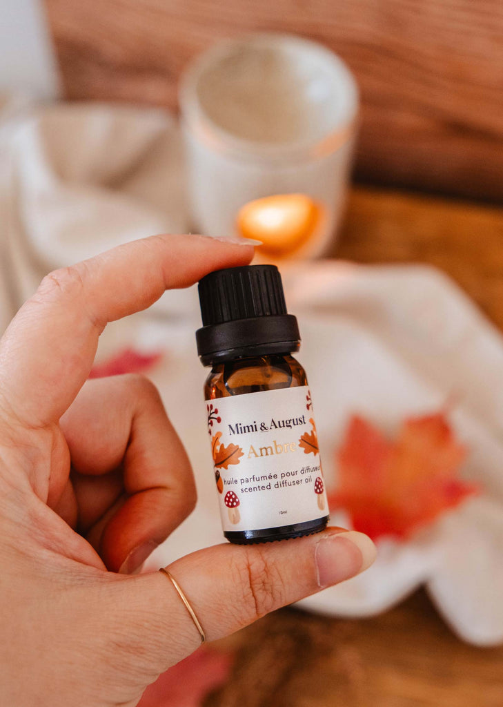 A hand holds a small bottle labeled "Scented Oil - Ambre" by Mimi & August, with the tropical foliage of a diffuser in the background, hinting at a luxurious Amalfi scent.