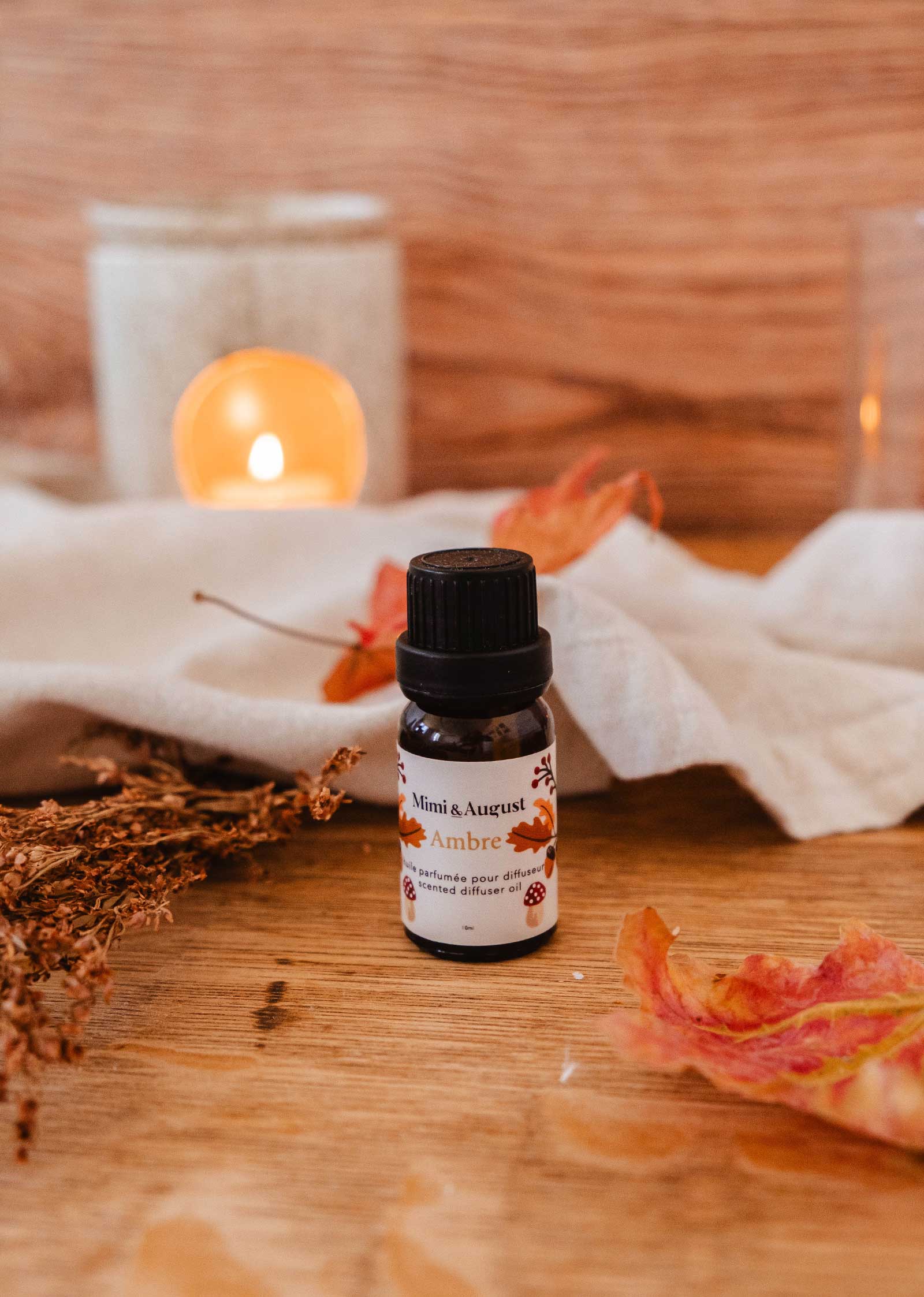 A small bottle labeled "Scented Oil - Ambre" by Mimi & August, perfect for use in a diffuser, is placed on a wooden surface with a lit candle, fabric, and dried leaves in the background.